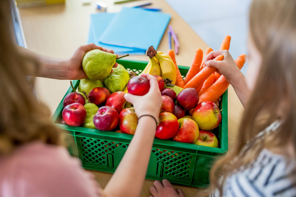 Kinder greifen in eine grüne Ökokiste voller Obst und Gemüse, die auf einem Schulpult steht
