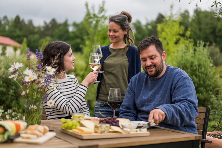 Drei Personen genießen draußen Wein und eine Käseplatte an einem Tisch.