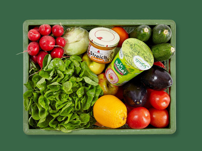 KI generiert: Das Bild zeigt einen grünen Korb voller frischer Lebensmittel, darunter Salat, Radieschen, Zitronen und Auberginen. In der Mitte befinden sich zwei Behälter mit Aufstrichen oder Sojaprodukten.