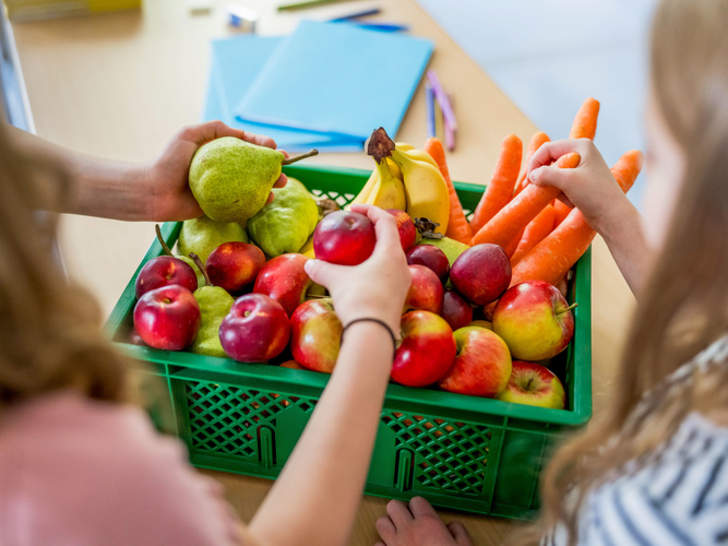 Kinder greifen in eine grüne Ökokiste voller Obst und Gemüse, die auf einem Schulpult steht