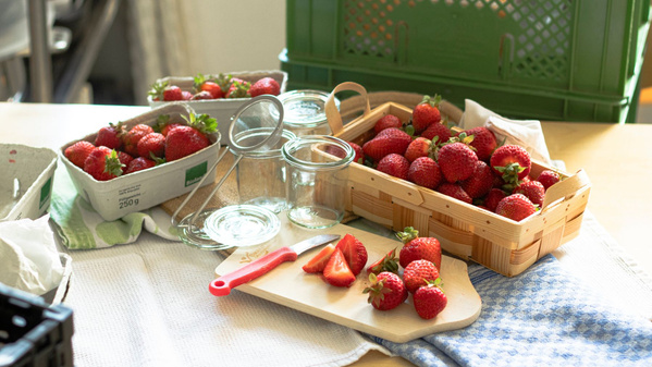 KI generiert: Frische Erdbeeren in Körben mit einem Schneidebrett, Glasgefäßen und einem Messer auf einem Tisch.