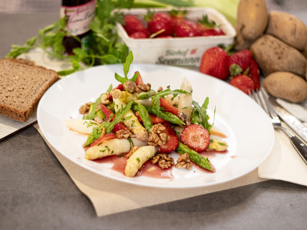 KI generiert: Ein Teller mit Spargel-Erdbeer-Salat und Walnüssen. Frische Erdbeeren und Kartoffeln im Hintergrund.