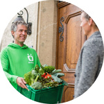 Mann reicht einer Frau an der Haustür eine Ökokiste mit firschem Obst und Gemüse.