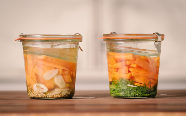 KI generiert: Zwei Einmachgläser mit eingelegtem Gemüse und Kräutern auf einem Tisch.