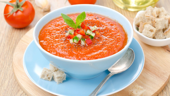 KI generiert: Ein Teller Gazpacho, garniert mit Gemüsewürfeln und Kräutern, auf einem Tisch mit Brotstücken und Tomaten.