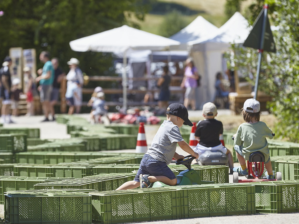 KI generiert: Kinder fahren auf Spielzeugfahrzeugen durch einen Parcours aus grünen Kisten bei einer Outdoor-Veranstaltung.